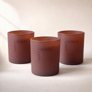 Three matte chestnut glass candle holders with the brand name 'KISLEV' printed vertically, arranged on a warm beige surface.