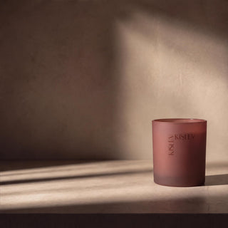 A matte glass candle jar in a muted chestnut color, standing on a surface with dramatic diagonal sunlight and shadow casting across the background. The brand name 'KISLEY' is printed vertically on the front of the jar.