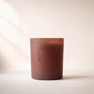 A matte glass candle jar in a warm chestnut brown color, featuring the brand name 'KISLEV' vertically on the front, set against a minimalist beige background with soft diagonal light shadows.