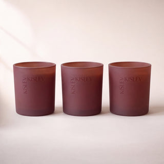 Three glass candle holders in a chestnut brown color, arranged in a row on a light beige background. Each holder features the brand name 'KISLEV' printed vertically in a matching tone.