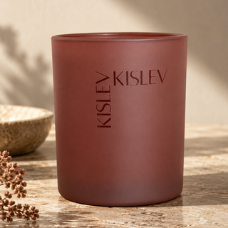 A matte glass candle jar in a muted chestnut brown color, standing on a stone surface with dried flowers and a bowl in the background. The brand name 'KISLEV' is printed vertically on the front.