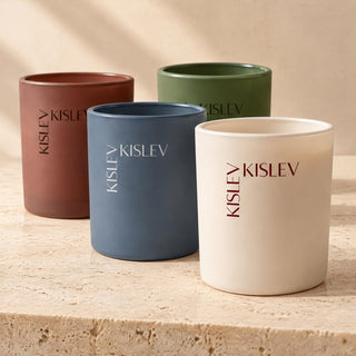 Four glass candles in matte colors—chestnut, ocean blue, willow green, and linen—arranged on a light stone surface. Each candle has the brand name 'KISLEV' printed vertically on the side.