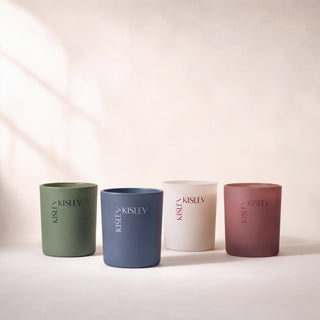 Four matte glass candle jars in muted willow green, ocean blue, linen, and chestnut, arranged in a row on a light beige surface with soft shadows. Each jar is printed with the brand name 'KISLEV' vertically.
