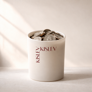 A linen glass jar filled with coins, featuring the name 'KISLEV' printed vertically in dark brown on the front, set against a warm beige background with soft diagonal shadows.