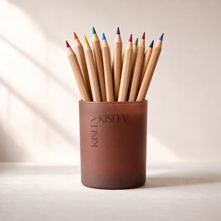 A chestnut brown glass jar used as a pencil holder with the brand name 'KISLEV' embossed on the front, filled with wooden colored pencils of various colors, set against a warm beige background with soft diagonal shadows.
