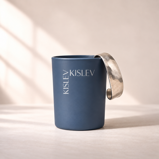 An ocean blue glass jar with a wide, hammered silver metal cuff displayed, featuring the name 'KISLEV' printed vertically on the front. The container is set against a warm beige background with soft window shadow patterns.