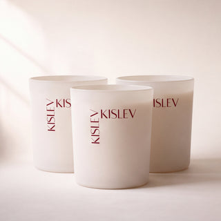 Three matte linen glass candle jars with the brand name 'KISLEV' printed vertically in dark brown on the front, arranged on a warm cream surface with soft diagonal light rays.