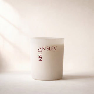 A matte glass candle jar with the brand name 'KISLEV' printed vertically in dark brown on the front, set against a warm beige background with soft diagonal shadows.