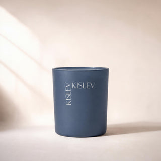 A matte ocean blue cylindrical candle jar with the brand name 'KISLEV' printed vertically on the front, set against a warm beige background with soft diagonal shadows.