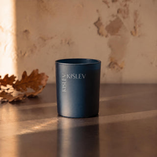 An ocean blue cylindrical candle jar with 'KISLEV' printed vertically on the front, set on a reflective wooden surface against a textured beige wall with warm sunlight and a dried leaf.