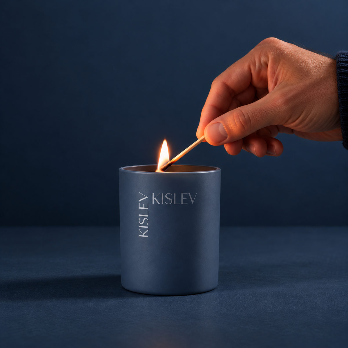 A hand holding a lit match above a matte oceanblue candle jar printed with the brand name KISLEV, set against a dark blue background.