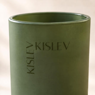 A close-up of a matte osage green cylindrical candle with the brand name 'KISLEV' printed vertically and horizontally on the front, set against a soft, warm beige background.