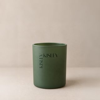 A matte sage green glass jar candle with the brand name 'KISLEV' printed vertically on the front, set against a neutral beige background.